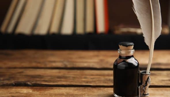 A glass ink bottle with a cork stopper and a quill pen resting in a holder on a wooden table. - Olive Oil Times