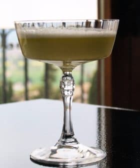 A crystal glass filled with a light green cocktail placed on a reflective surface. - Olive Oil Times