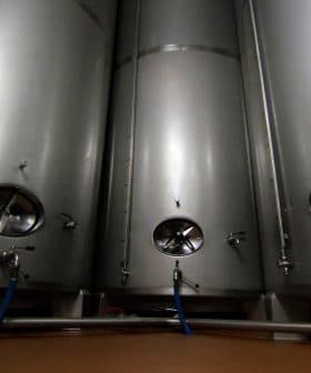 Three stainless steel fermentation tanks with circular openings and valves in a dimly lit space. - Olive Oil Times