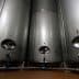 Three stainless steel fermentation tanks with circular openings and valves in a dimly lit space. - Olive Oil Times