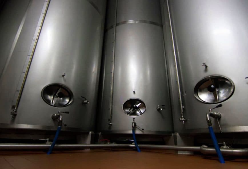 Three stainless steel fermentation tanks with circular openings and valves in a dimly lit space. - Olive Oil Times