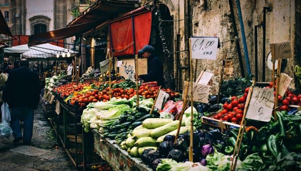 Market stall displaying various vegetables and fruits with price signs in a bustling market setting. - Olive Oil Times