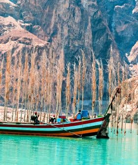 A colorful fishing boat anchored in turquoise water with tall, bare trees in the background. - Olive Oil Times