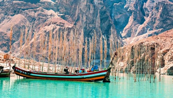 A colorful fishing boat anchored in turquoise water with tall, bare trees in the background. - Olive Oil Times