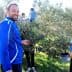 Group of people participating in olive harvesting, with one man smiling and others collecting olives from trees. - Olive Oil Times