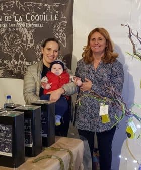 Two women and a baby at an olive oil stand with products displayed in a tent. - Olive Oil Times