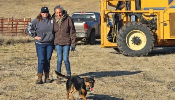 Two women standing next to a yellow machine with a dog running in front of them in an open field. - Olive Oil Times