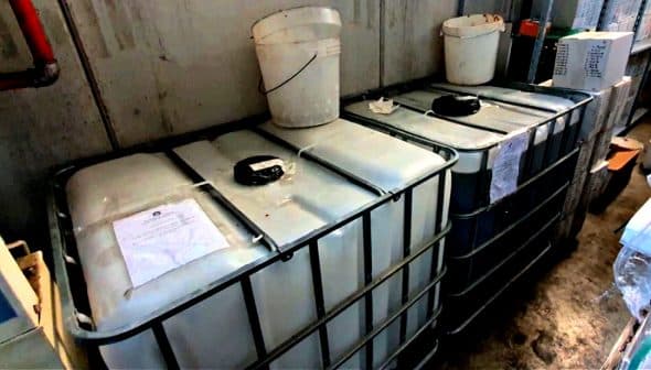 Two large industrial storage tanks with a white bucket placed on top in a warehouse setting. - Olive Oil Times