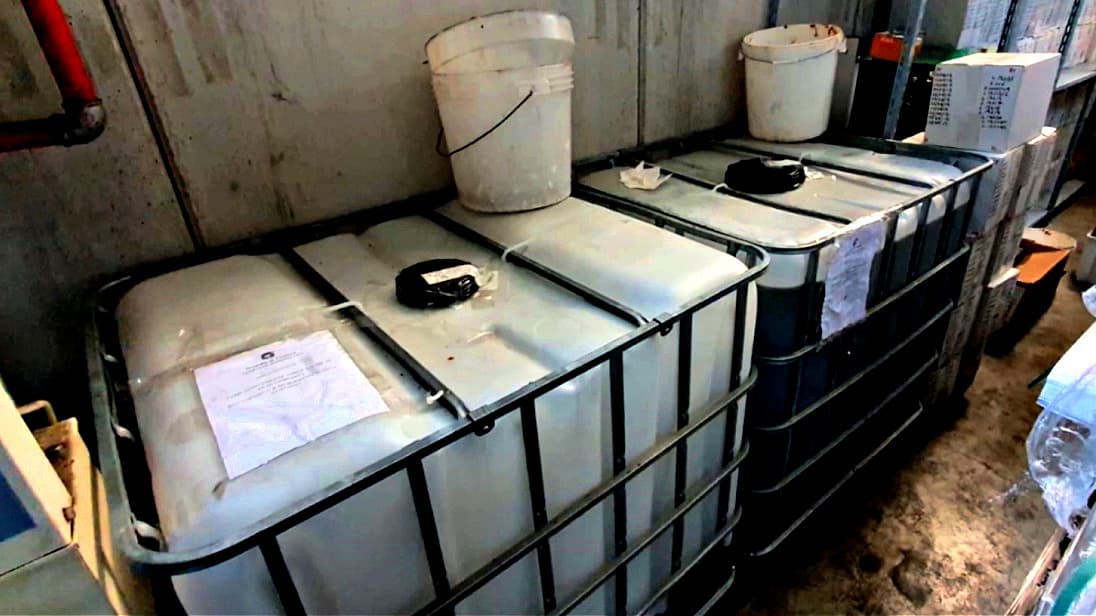 Two large industrial storage tanks with a white bucket placed on top in a warehouse setting. - Olive Oil Times