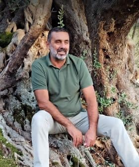 Man with a beard sitting on the roots of an olive tree, wearing a green shirt and light-colored pants. - Olive Oil Times