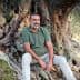 Man with a beard sitting on the roots of an olive tree, wearing a green shirt and light-colored pants. - Olive Oil Times