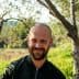 Man with a beard smiling while standing between olive trees in a natural setting. - Olive Oil Times