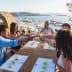 A group of five people toasting with drinks at a wooden table overlooking a coastal view. - Olive Oil Times