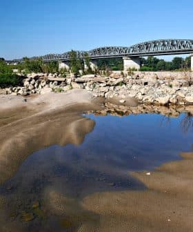 A metal bridge spans a riverbank with sandy terrain and a small pool of water in the foreground. - Olive Oil Times