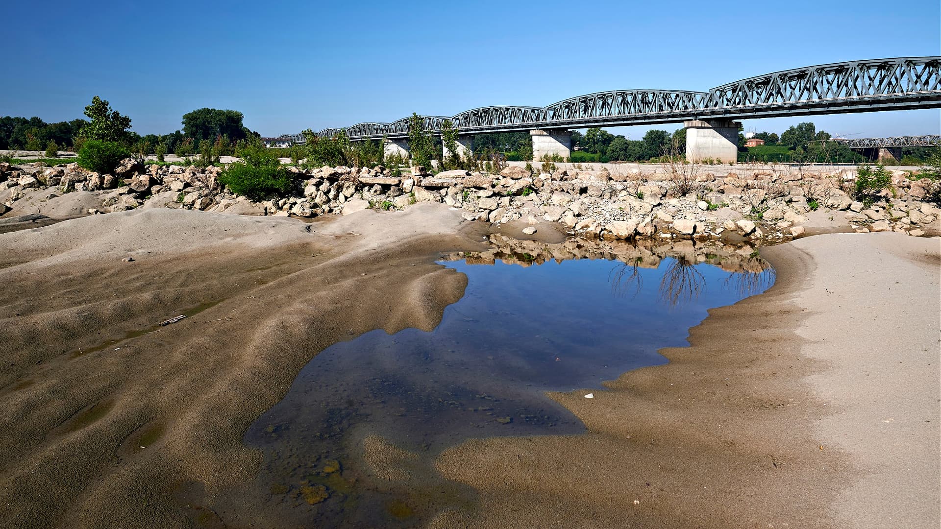 A metal bridge spans a riverbank with sandy terrain and a small pool of water in the foreground. - Olive Oil Times