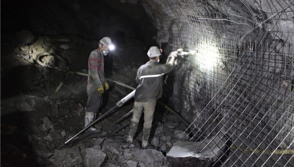 Two miners working in a dark underground mine with safety gear and tools. - Olive Oil Times