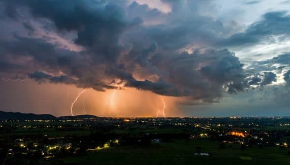 Lightning illuminating the sky over a landscape during dusk with dark clouds and distant lights. - Olive Oil Times