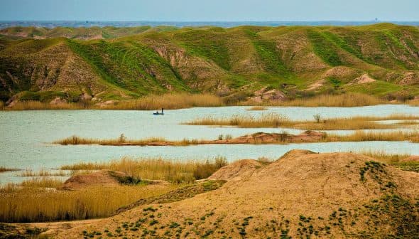 A serene landscape featuring green hills, a water body, and a small boat in the distance. - Olive Oil Times