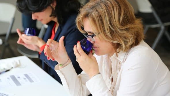 Two women participating in an olive oil tasting session, holding blue glasses and examining the aroma. - Olive Oil Times