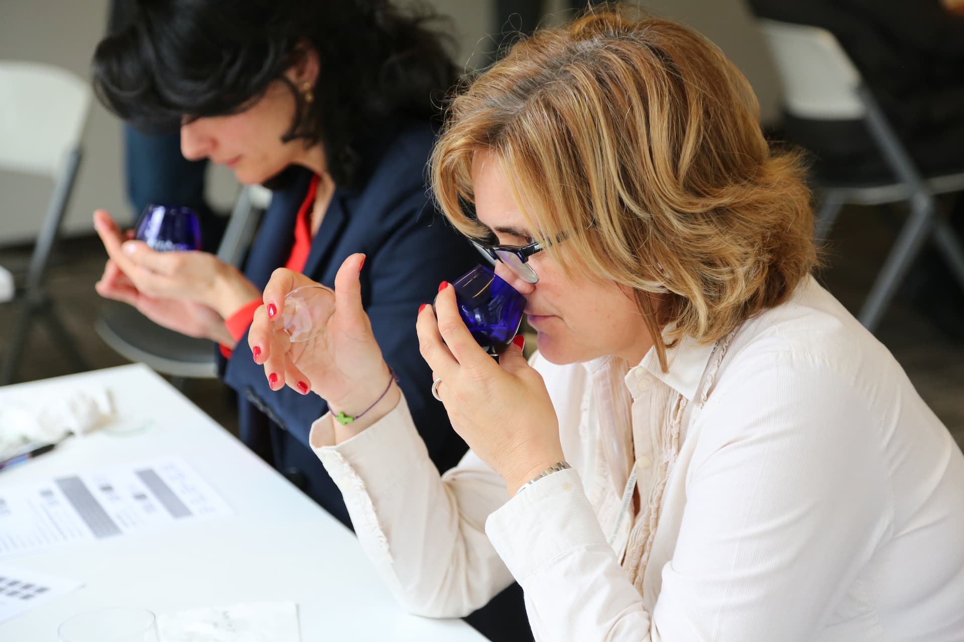 Two women participating in an olive oil tasting session, holding blue glasses and examining the aroma. - Olive Oil Times