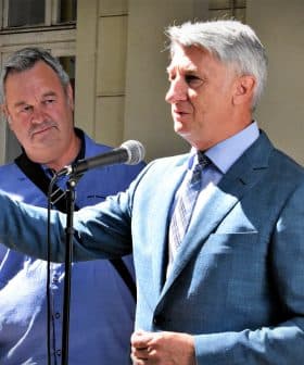 Two men, one speaking into a microphone while the other listens attentively, standing outdoors. - Olive Oil Times