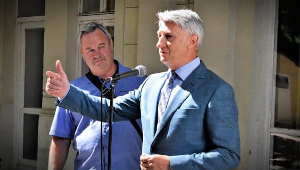 Two men, one speaking into a microphone while the other listens attentively, standing outdoors. - Olive Oil Times