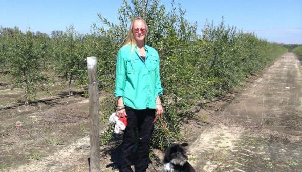 Woman wearing a green shirt and black pants standing with a black dog in an olive grove. - Olive Oil Times
