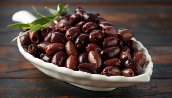 A white bowl filled with a variety of dark olives, garnished with a sprig of olive leaves. - Olive Oil Times