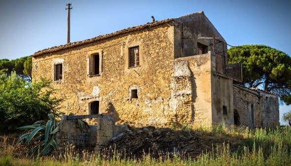 Abandoned stone house with a rustic exterior and overgrown vegetation in a rural area. - Olive Oil Times