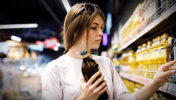 Woman with brown hair holding a bottle of olive oil while examining products on a shelf in a grocery store. - Olive Oil Times