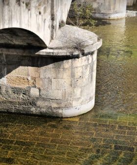 Close-up view of a stone bridge arching over a calm body of water with visible textures and reflections. - Olive Oil Times