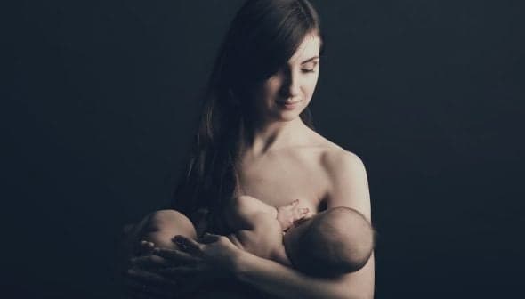 A mother gently cradling her baby in a dark background, both appearing calm and serene. - Olive Oil Times