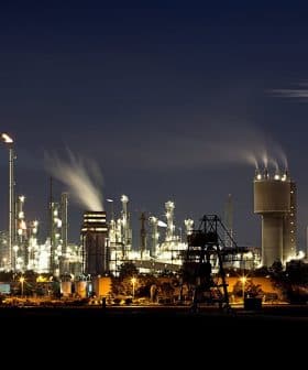 Night view of an industrial oil refinery with illuminated structures and smoke emissions from chimneys. - Olive Oil Times