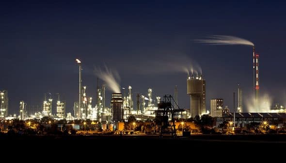 Night view of an industrial oil refinery with illuminated structures and smoke emissions from chimneys. - Olive Oil Times