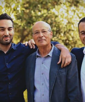 Three men standing outdoors, one with an arm around another, all smiling at the camera. - Olive Oil Times