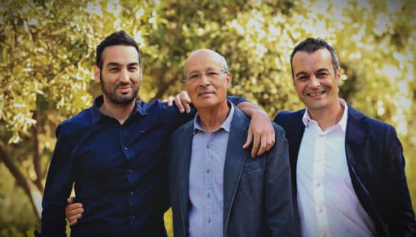 Three men standing outdoors, one with an arm around another, all smiling at the camera. - Olive Oil Times