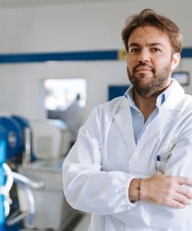 Man wearing a laboratory coat standing with arms crossed in a facility with machinery in the background. - Olive Oil Times