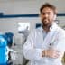 Man wearing a laboratory coat standing with arms crossed in a facility with machinery in the background. - Olive Oil Times