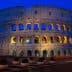 The Colosseum in Rome illuminated at dusk with a blue sky backdrop and lit arches. - Olive Oil Times