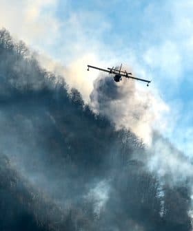 An aircraft releasing water to combat a wildfire in a mountainous region with smoke rising from the ground. - Olive Oil Times