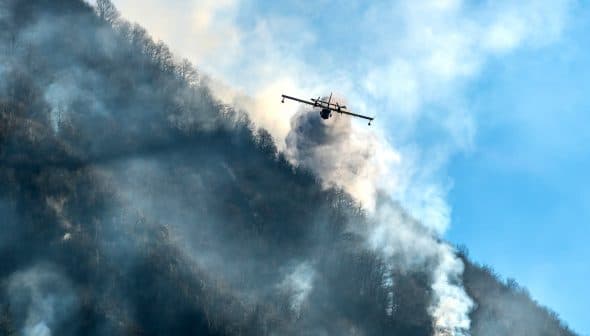 An aircraft releasing water to combat a wildfire in a mountainous region with smoke rising from the ground. - Olive Oil Times