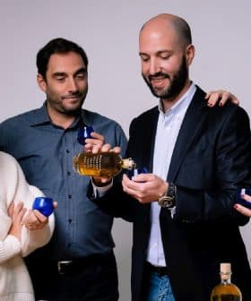 Four individuals holding olive oil bottles and small blue cups in a neutral setting. - Olive Oil Times