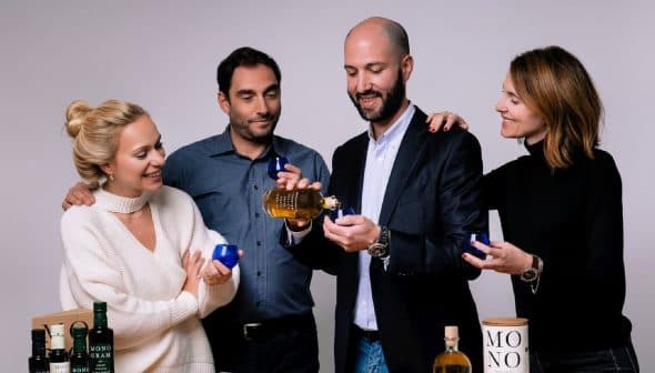 Four individuals holding olive oil bottles and small blue cups in a neutral setting. - Olive Oil Times
