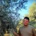 A man standing next to olive trees in an outdoor setting, with green foliage in the background. - Olive Oil Times