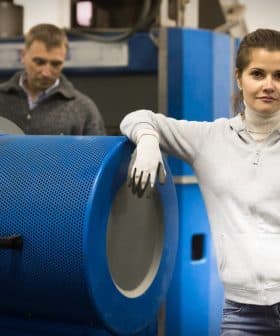 Woman in a gray sweater and gloves standing next to industrial machinery in a factory setting. - Olive Oil Times