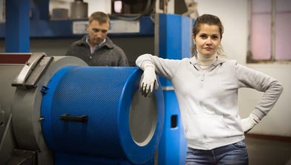 Woman in a gray sweater and gloves standing next to industrial machinery in a factory setting. - Olive Oil Times