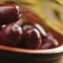 A close-up view of dark olives in a brown bowl with olive branches in the background. - Olive Oil Times