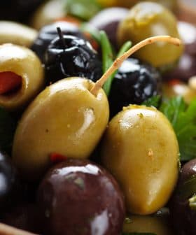 A close-up of mixed green and black olives garnished with herbs in a wooden bowl. - Olive Oil Times