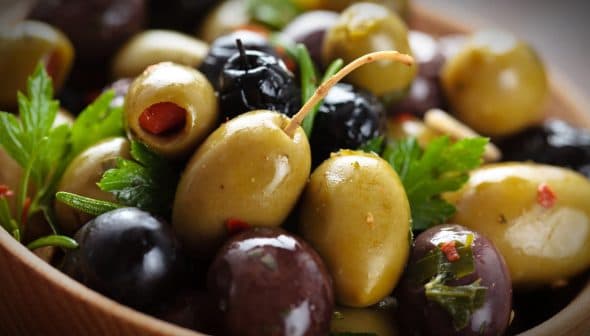 A close-up of mixed green and black olives garnished with herbs in a wooden bowl. - Olive Oil Times