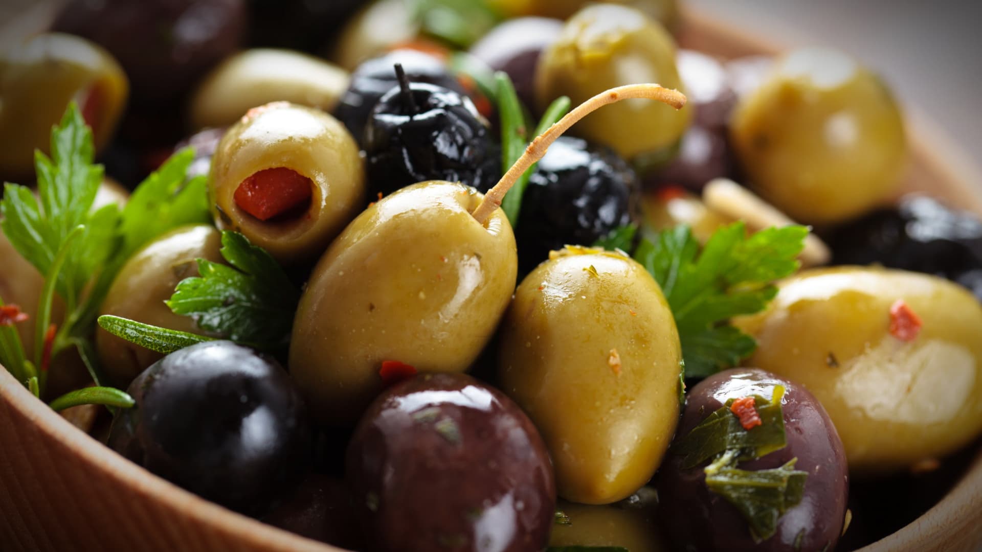 A close-up of mixed green and black olives garnished with herbs in a wooden bowl. - Olive Oil Times